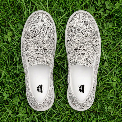 A pair of slip-on canvas shoes with intricate black and white doodle-style patterns, displayed from a top-down view on a grass background.